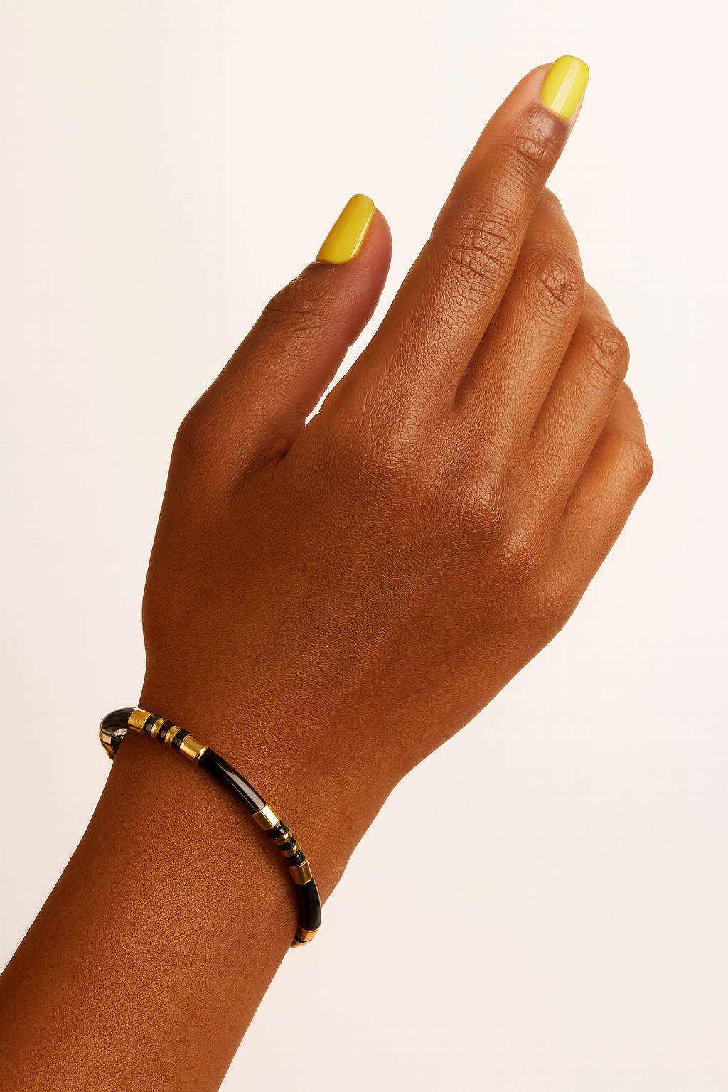 Hand with yellow nail polish wearing a black beaded bracelet on a beige background. Close-up of black enamel and gold segmented cuff bracelet.