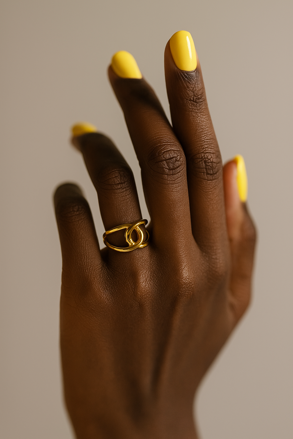 Hand with gold ring and yellow nail polish on a neutral background. Close-up of gold Bria Ring showcasing looped artistic structure.