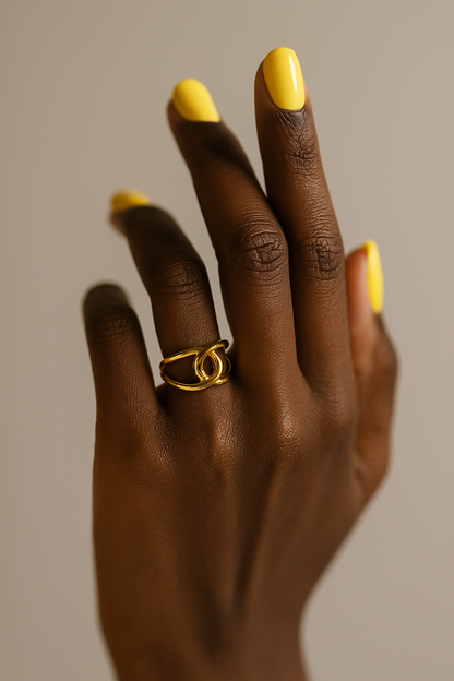 Hand with gold ring and yellow nail polish on a neutral background. Close-up of gold Bria Ring showcasing looped artistic structure.