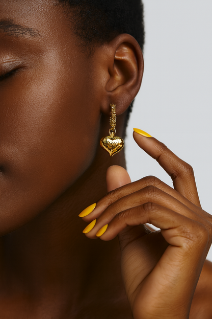 Close-up of a person wearing a gold heart-shaped earring with a neutral background. Pair of gold heart hoop earrings featuring quilt-texture heart charm. Lila Earrings  gold hoop with beaded detail and heart pendant