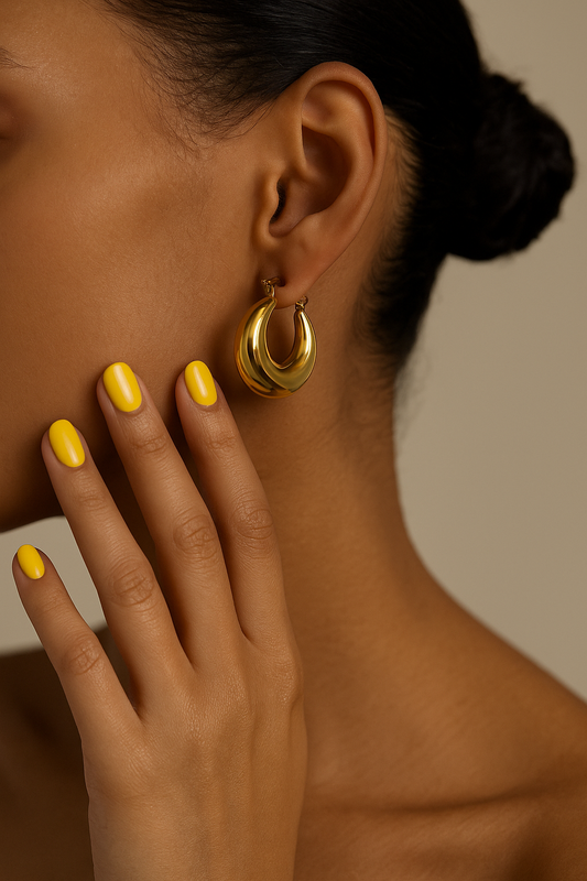 Close-up of a person wearing gold hoop earrings with yellow nail polish on a neutral background. High-shine gold hoop earrings with polished finish.