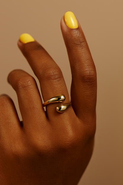 Hand wearing a gold ring with yellow nail polish on a brown background. Gold Sia Ring – sculptural modern open-ended ring with high-polish finish.
