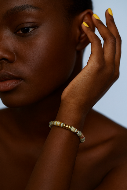 Woman with a gold and green beaded bracelet on her wrist, touching her ear against a neutral background.