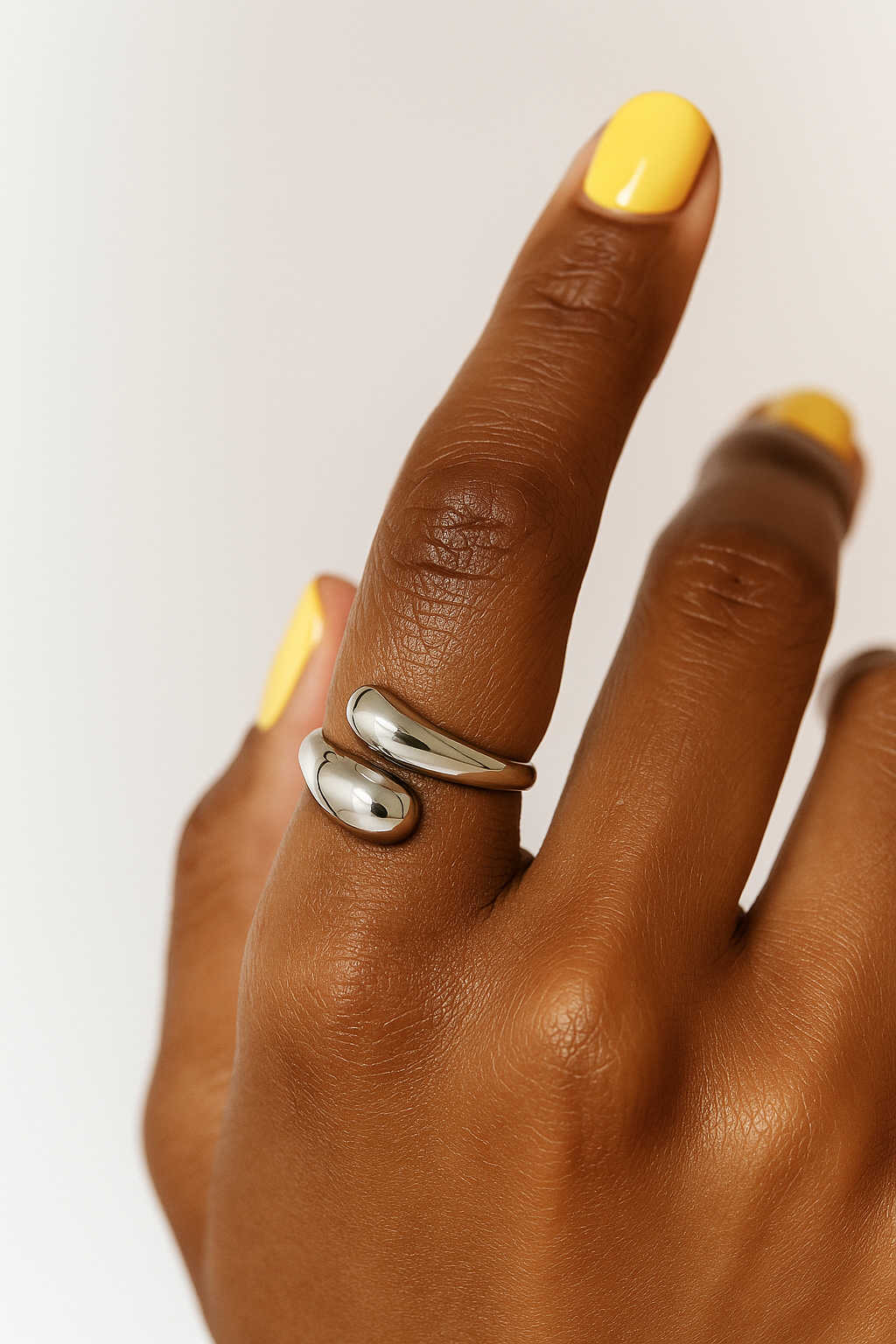 Hand wearing a silver ring with yellow nail polish on a white background.Silver Sia Ring – sculptural modern open-ended ring with high-polish finish.