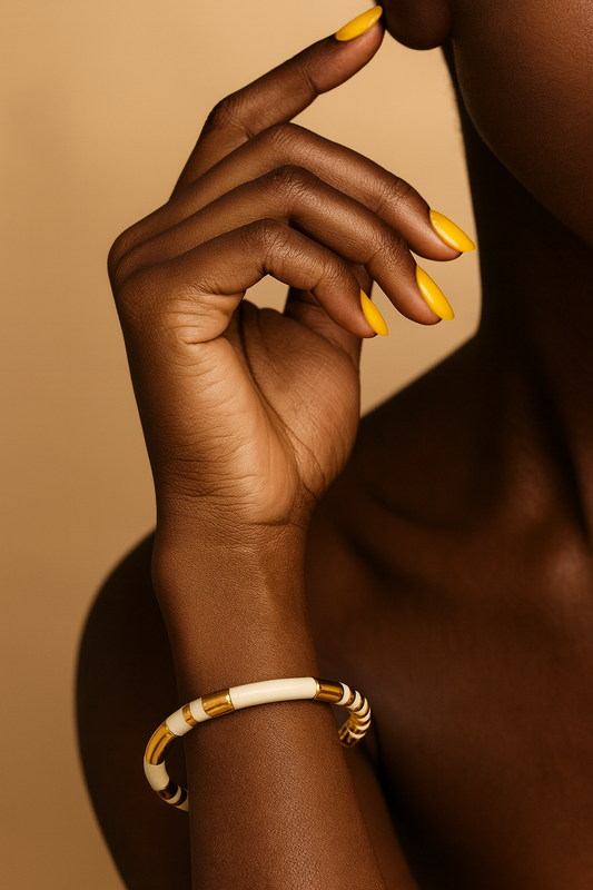 Close-up of white enamel and gold segmented cuff bracelet. Hand with yellow nail polish wearing a gold and white bracelet on a beige background